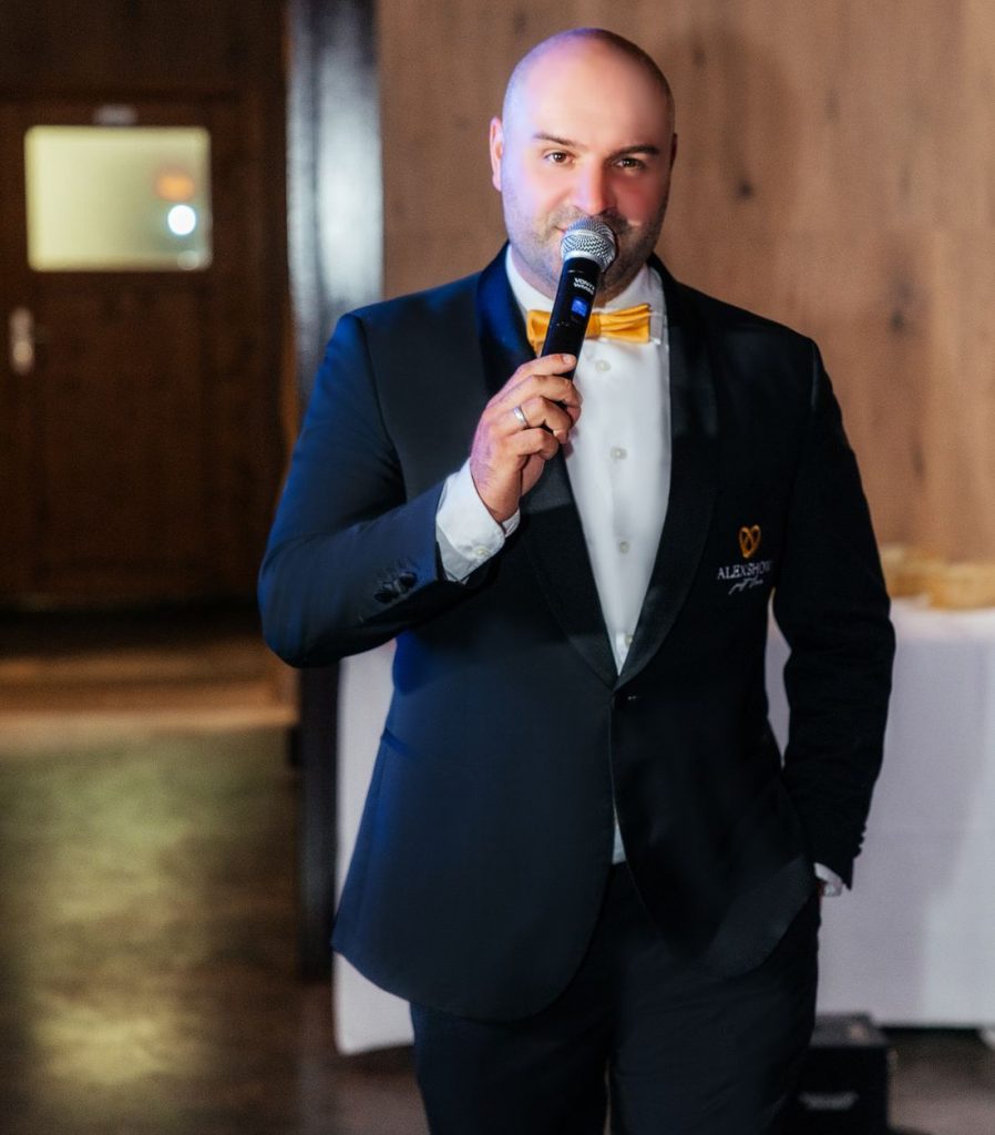 Hochzeitsmoderator Ivan von AlexShow im dunklen Anzug mit goldener Fliege spricht ins Mikrofon