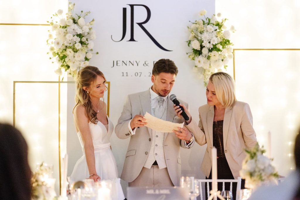 Hochzeitsmoderatorin Lera hält dem Bräutigam das Mikrofon, während er neben der Braut einen Text vorliest
