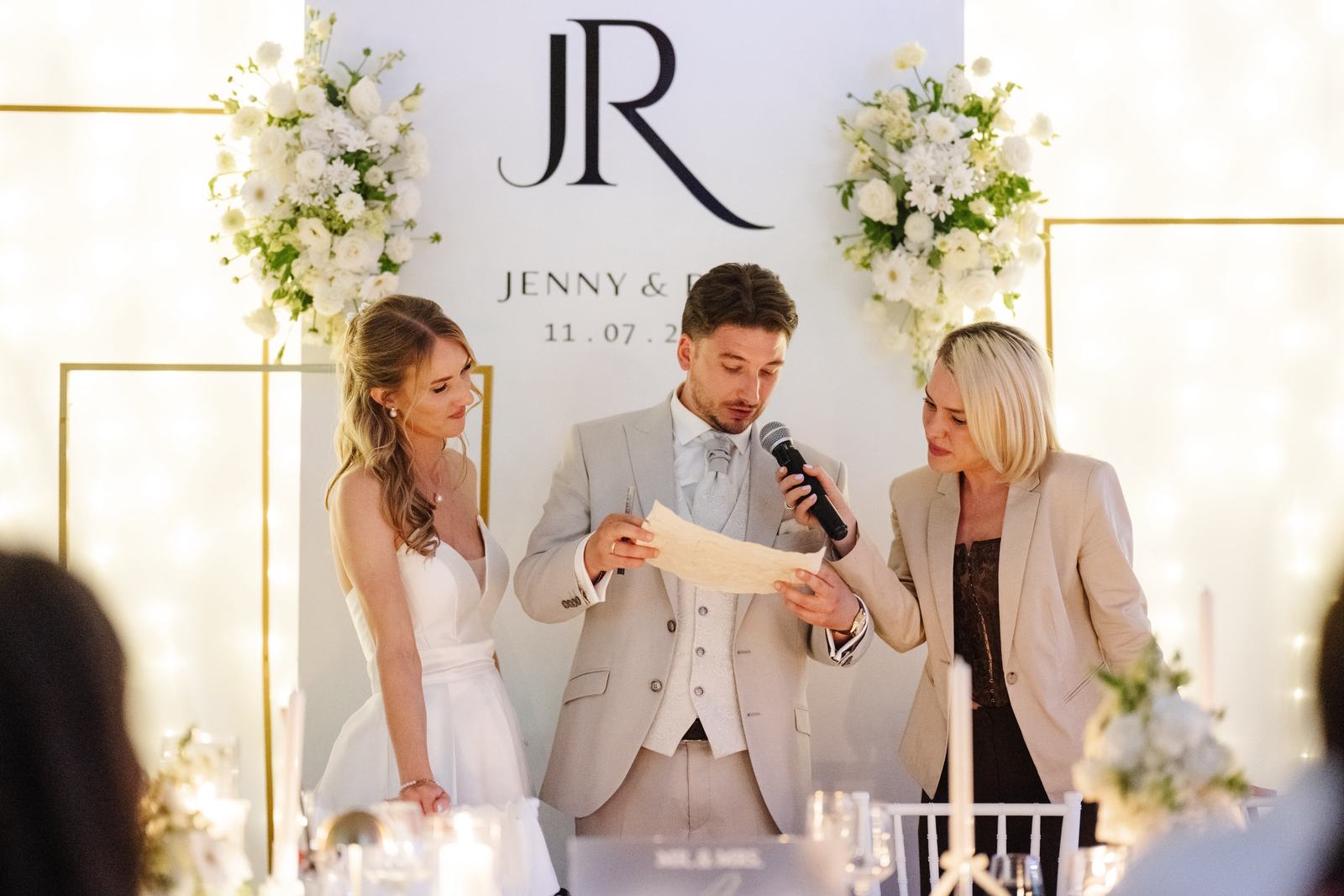 Hochzeitsmoderatorin Lera hält dem Bräutigam das Mikrofon, während er neben der Braut einen Text vorliest Hochzeitsmoderatorin Lera hält dem Bräutigam das Mikrofon, während er neben der Braut einen Text vorliest