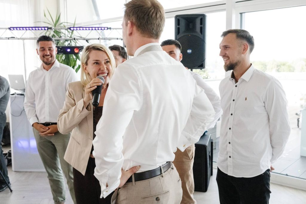 Hochzeitsmoderatorin Lera lacht mit dem Mikrofon in der Hand im Kreis der Hochzeitsgäste
