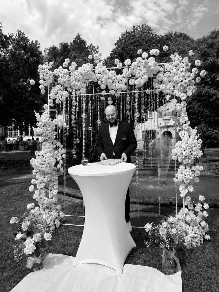 Hochzeitsmoderator Vincent von AlexShow bei einer freien Trauung – Schwarz-Weiß-Aufnahme vor der Blumenwand