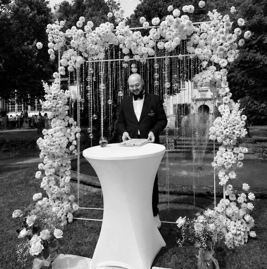 Hochzeitsmoderator Vincent von AlexShow bei einer freien Trauung – Schwarz-Weiß-Aufnahme vor der Blumenwand Hochzeitsmoderator Vincent von AlexShow bei einer freien Trauung – Schwarz-Weiß-Aufnahme vor der Blumenwand