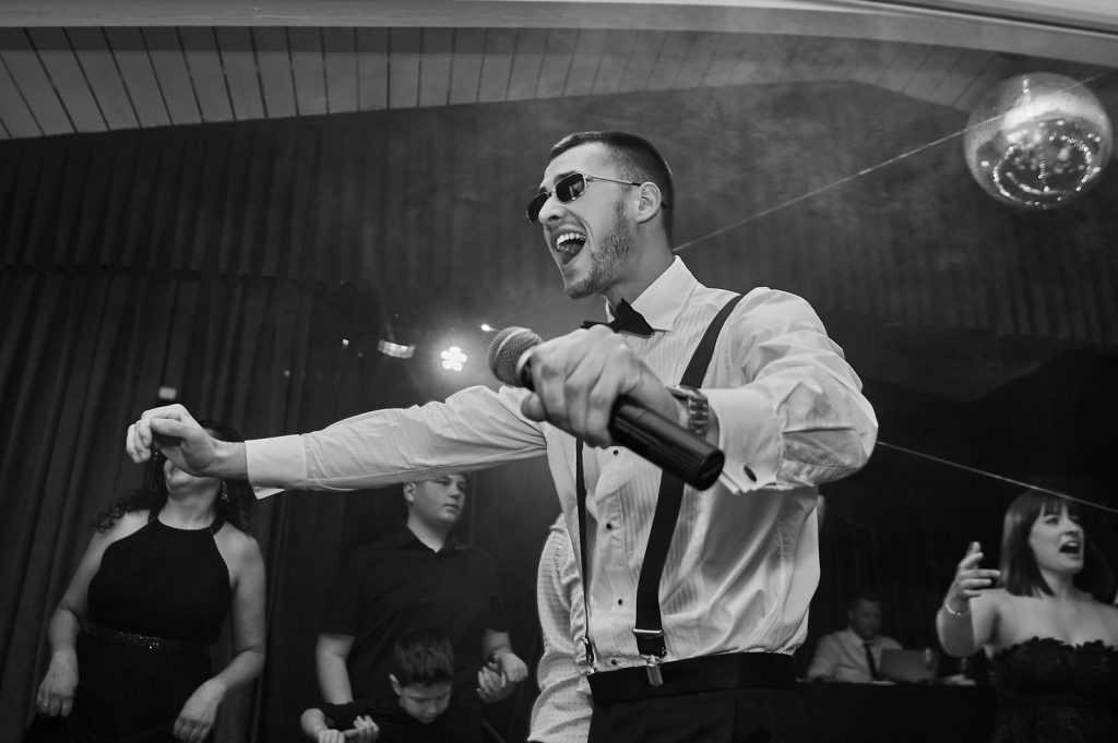 Hochzeitsmoderator Arti aus München mit Mikrofon und Hosenträgern moderiert energiegeladen die Hochzeitsparty, tanzende Gäste im Hintergrund