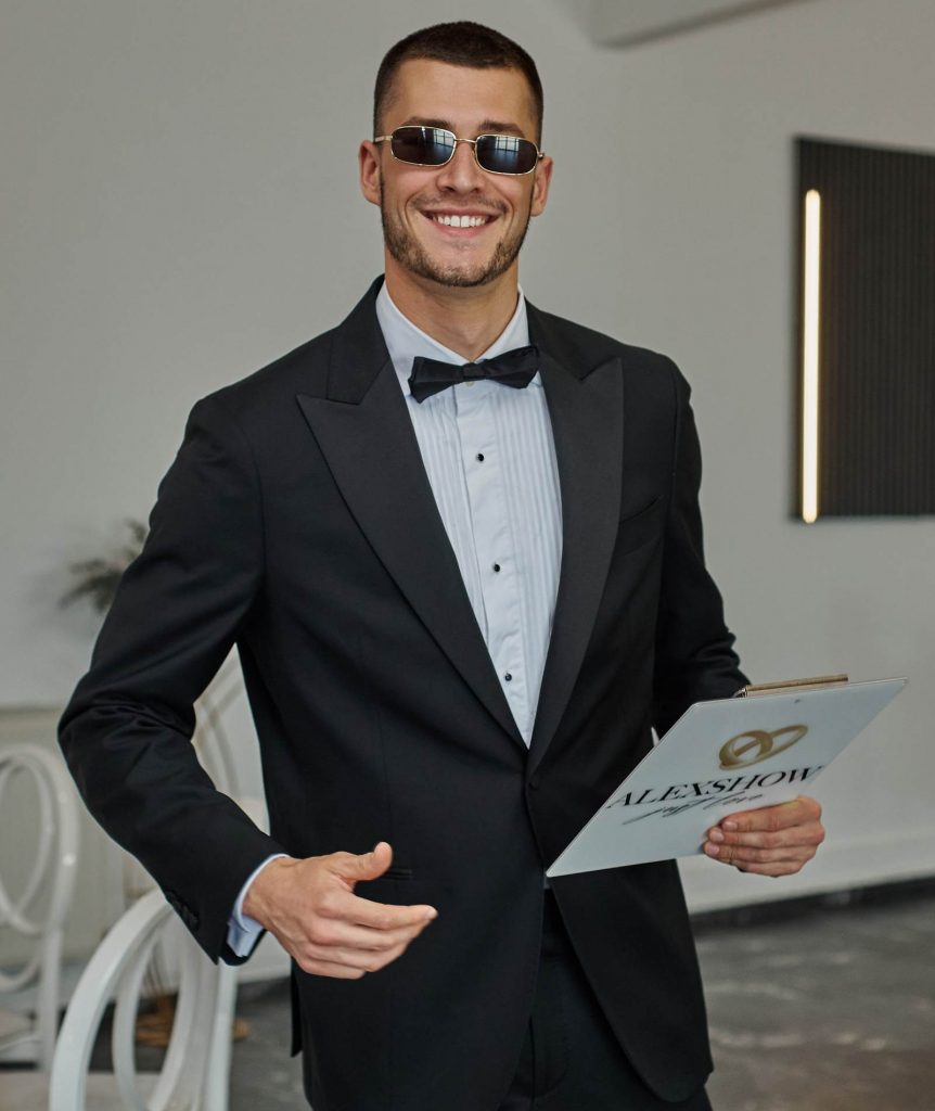 Hochzeitsmoderator Arti aus München im schwarzen Smoking mit Fliege und Sonnenbrille, lächelnd mit ALEXSHOW-Klemmbrett in der Hand