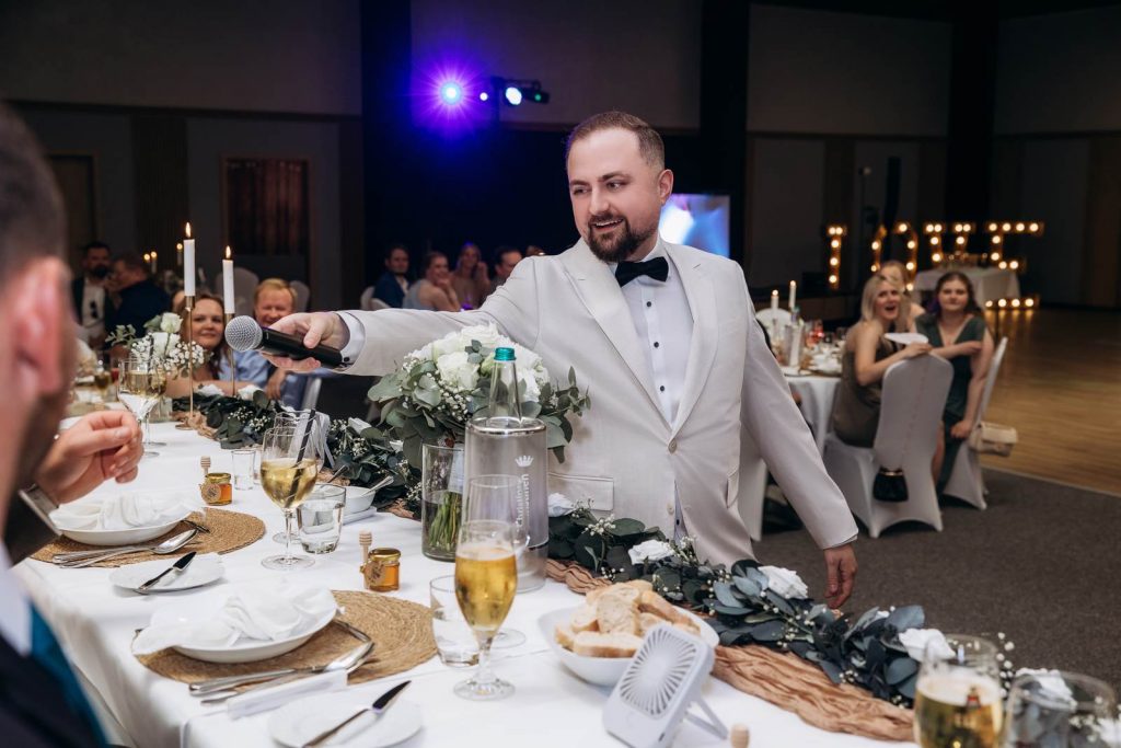 Hochzeitsmoderator Vladislav von AlexShow reicht bei der Hochzeitsfeier einem Gast das Mikrofon am festlich dekorierten Tisch