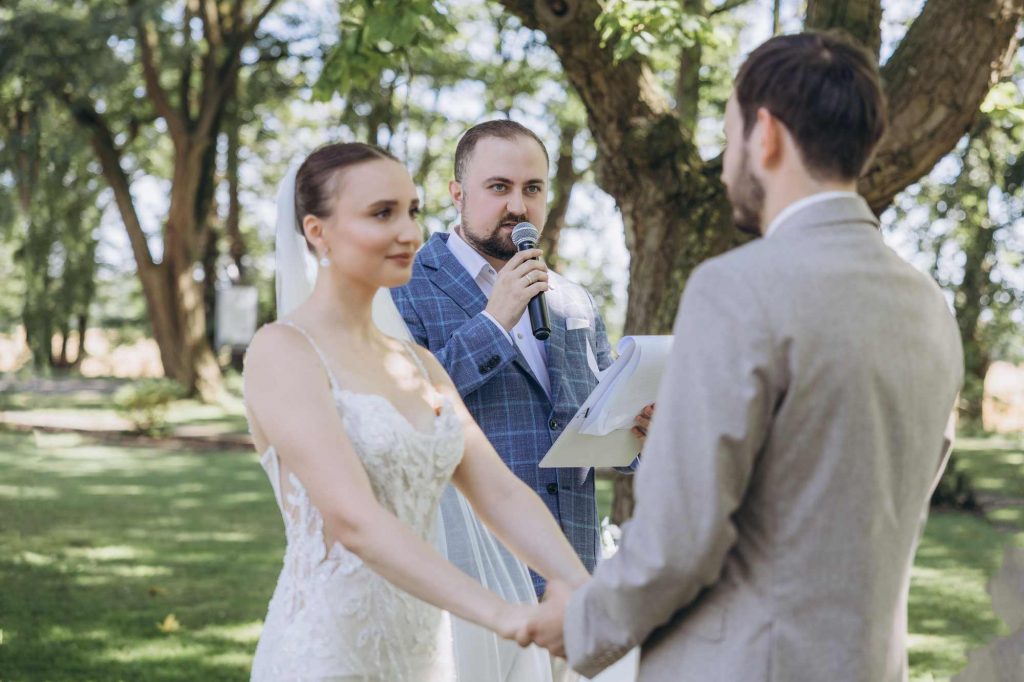 Hochzeitsmoderator Vladislav spricht ins Mikrofon zwischen Braut und Bräutigam beim Eheversprechen
