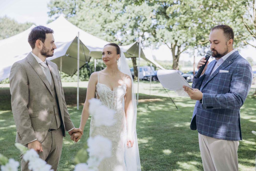 Hochzeitsmoderator Vladislav liest die Trauungsrede, Brautpaar hält Händchen vor Zeltdekoration