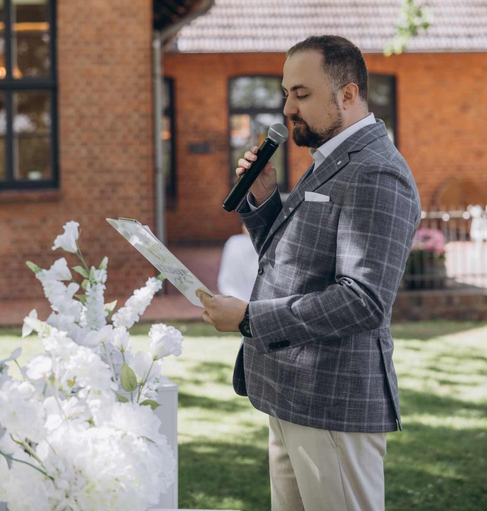 Hochzeitsmoderator Vladislav liest mit Mikrofon aus der Moderationsmappe vor einem Backsteinhaus