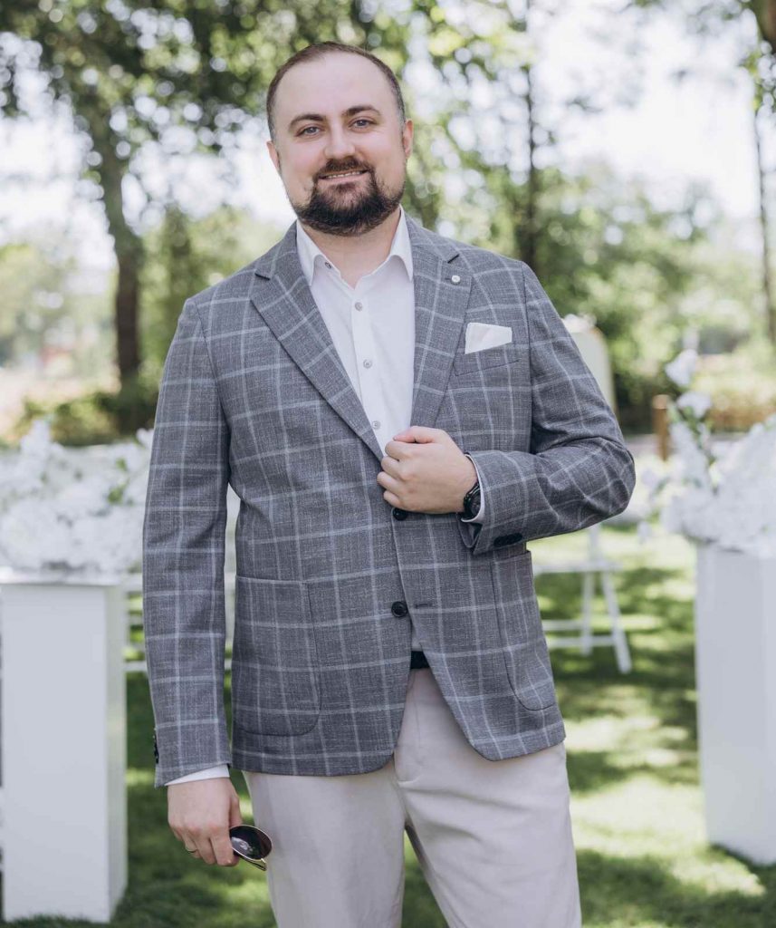 Hochzeitsmoderator Vladislav im grauen Karo-Sakko vor Blumendekoration im Garten