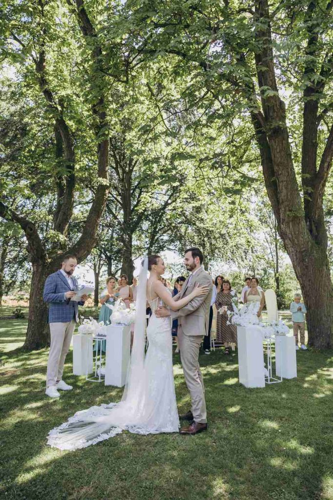 Brautpaar umarmt sich unter Bäumen, Hochzeitsmoderator Vladislav moderiert vor den Gästen