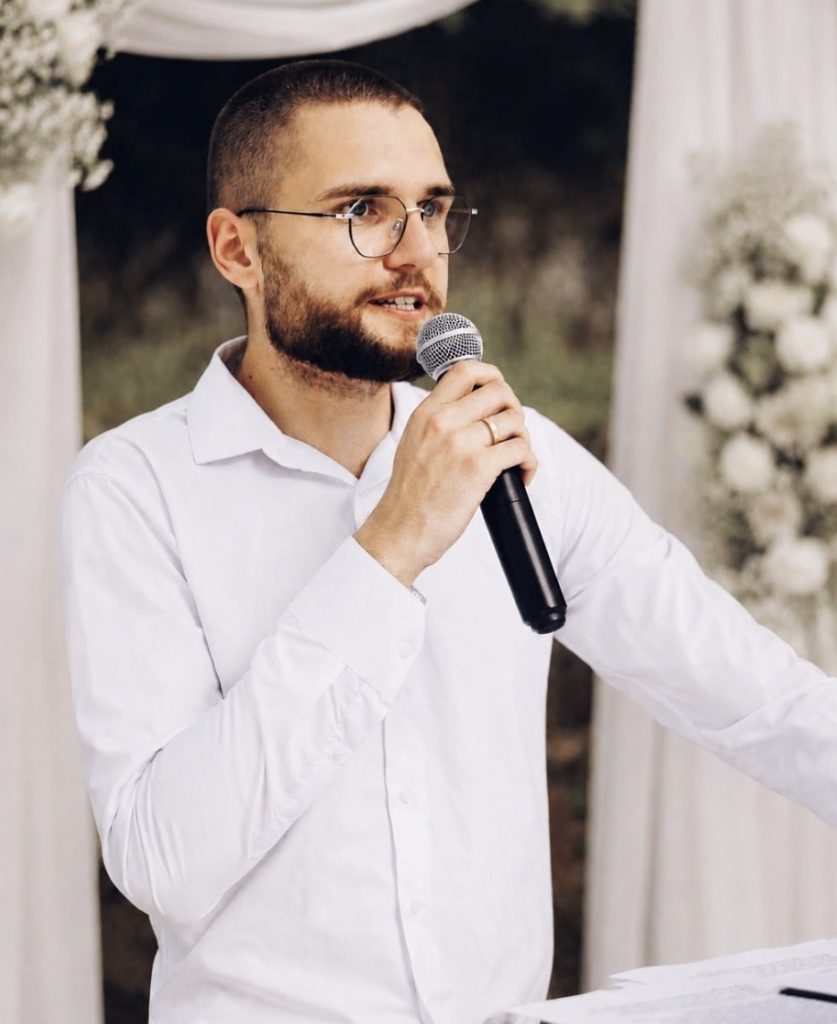 Hochzeitsmoderator Davis spricht mit Mikrofon vor einem weißen Blumenbogen bei einer freien Trauung