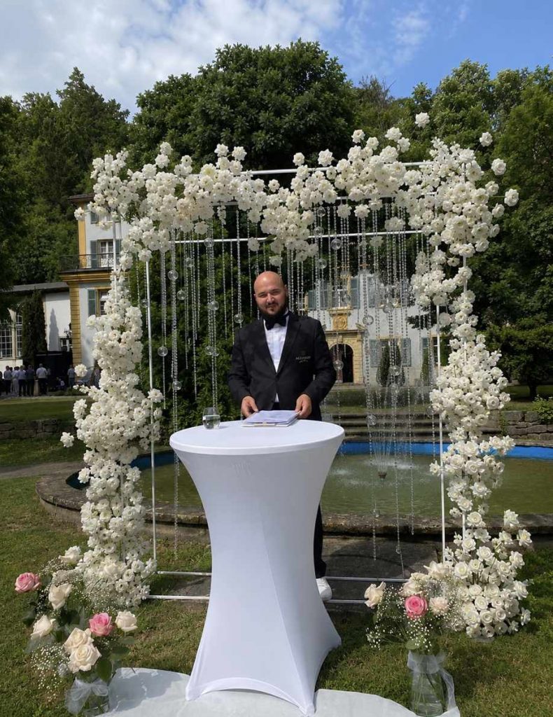 Hochzeitsmoderator Vincent von AlexShow bei einer freien Trauung vor einer weißen Blumenwand