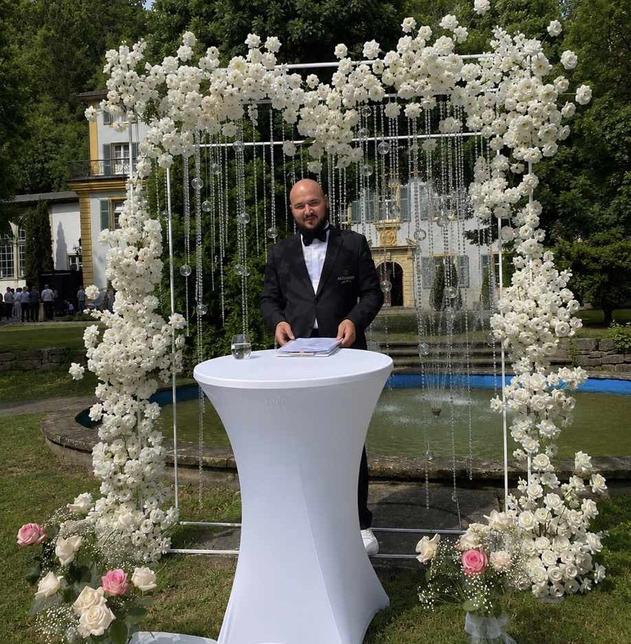 Hochzeitsmoderator Vincent von AlexShow bei einer freien Trauung vor einer weißen Blumenwand Hochzeitsmoderator Vincent von AlexShow bei einer freien Trauung vor einer weißen Blumenwand