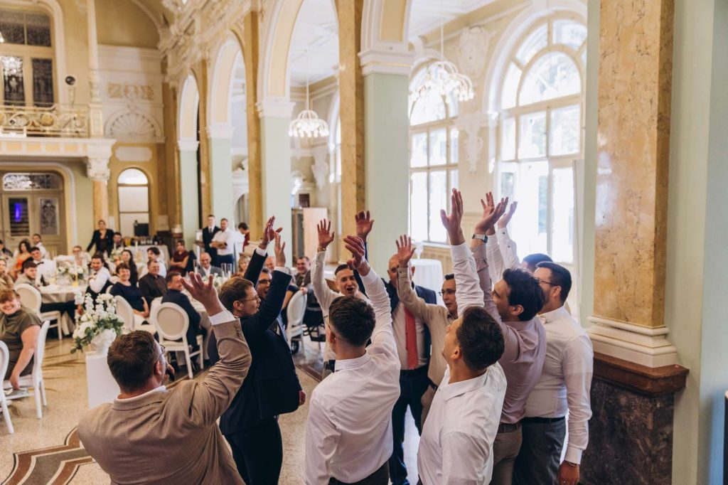Hochzeitsmoderator Demy feiert mit Gästen, die in einem festlichen Saal die Hände heben