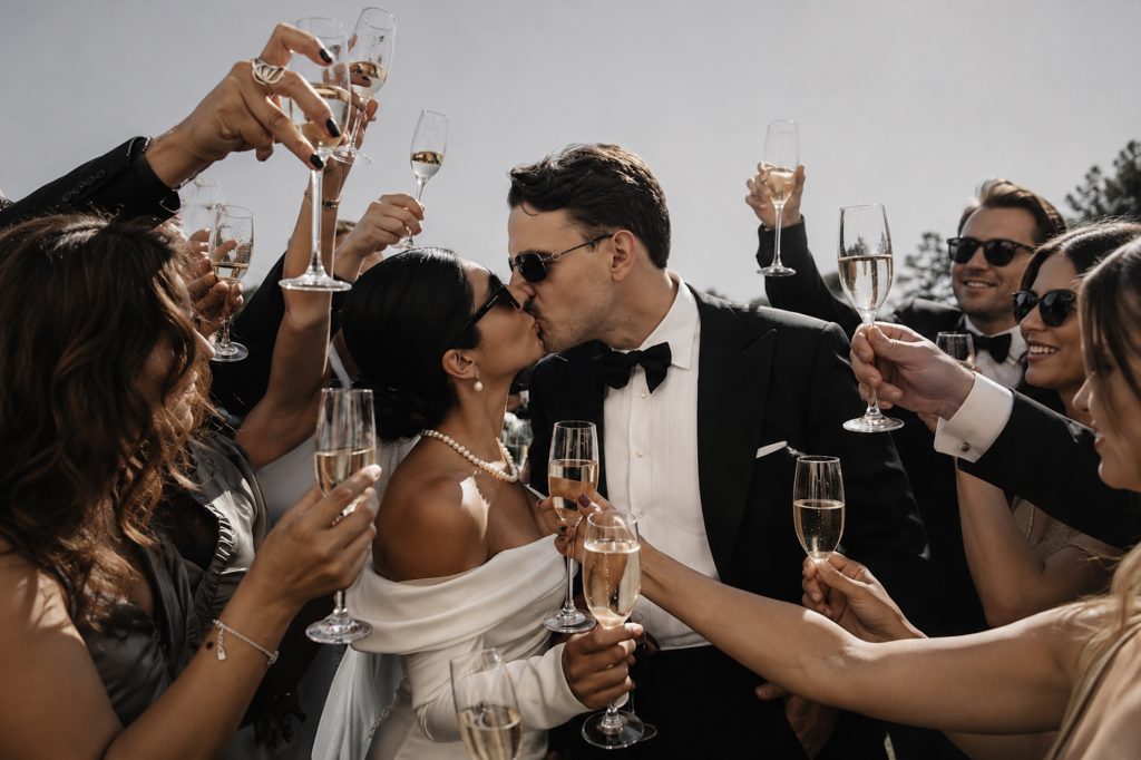 Braut und Bräutigam küssen sich beim Sektempfang auf einer russisch-französischen Hochzeit, während die Gäste mit Champagner anstoßen.