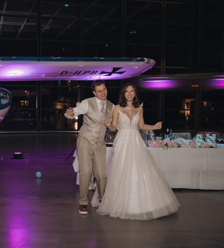 Brautpaar betritt lächelnd die Tanzfläche vor einem violett beleuchteten Flugzeug im Hangar