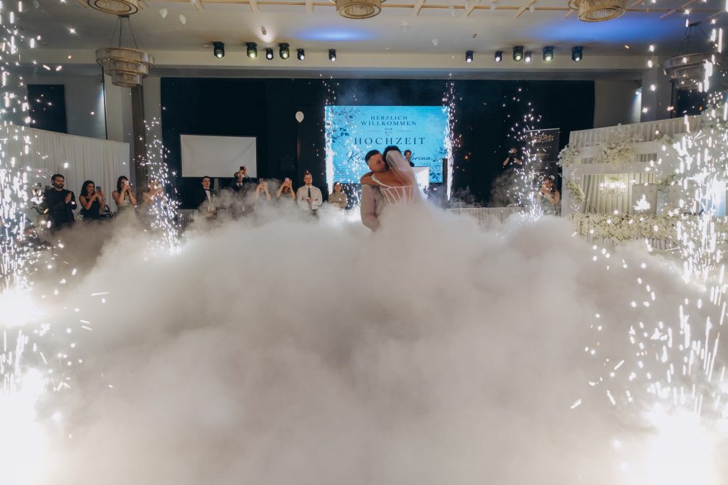 Brautpaar tanzt inmitten einer großen Nebelwolke und Funkenfontänen vor der Willkommens-Leinwand bei einer Hochzeit in Köln