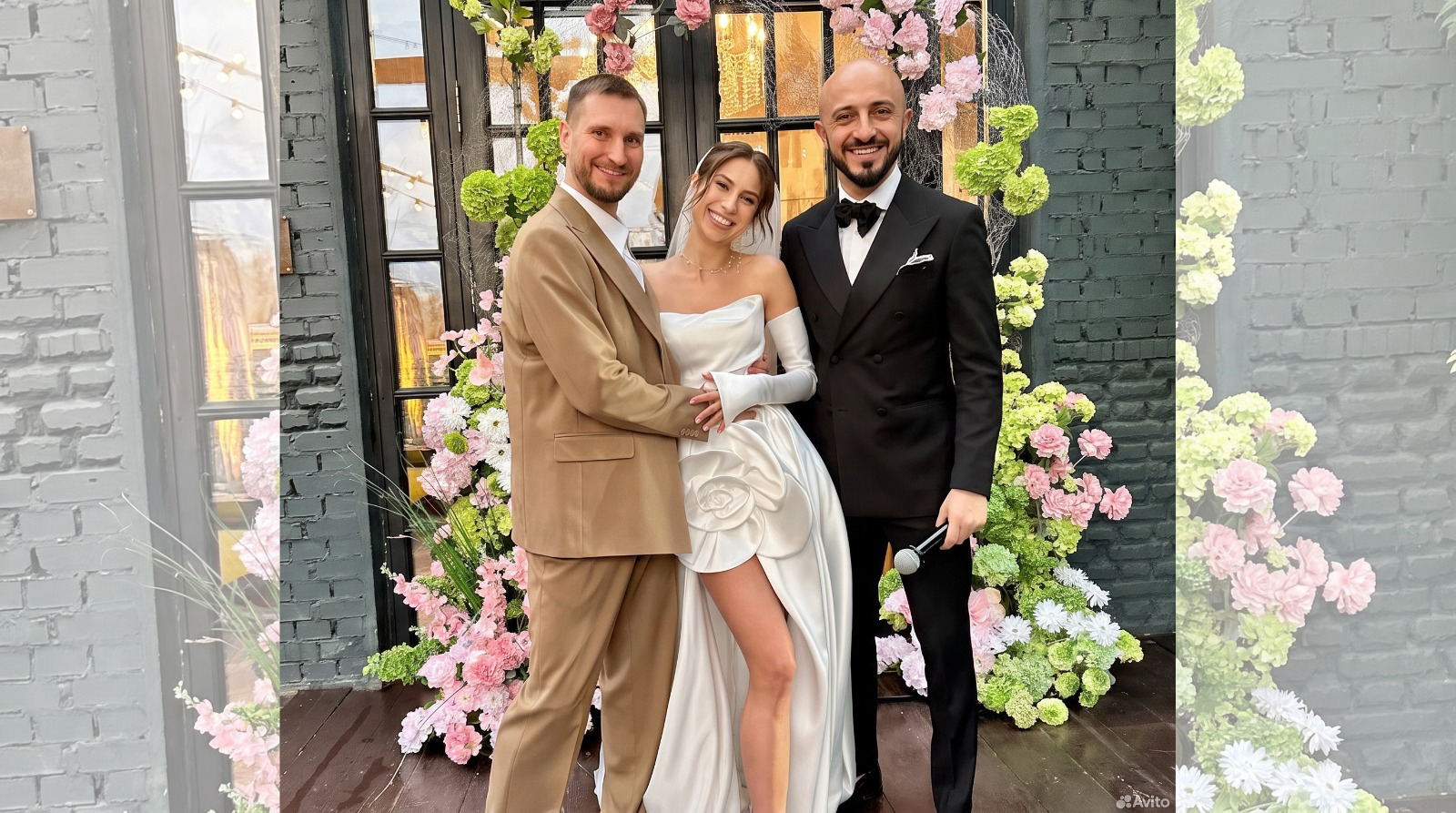 Jens mit dem Brautpaar vor Blumendekoration Jens im schwarzen Smoking mit Fliege und Mikrofon neben einem Brautpaar vor einer Tür mit rosa und grüner Blumendekoration.