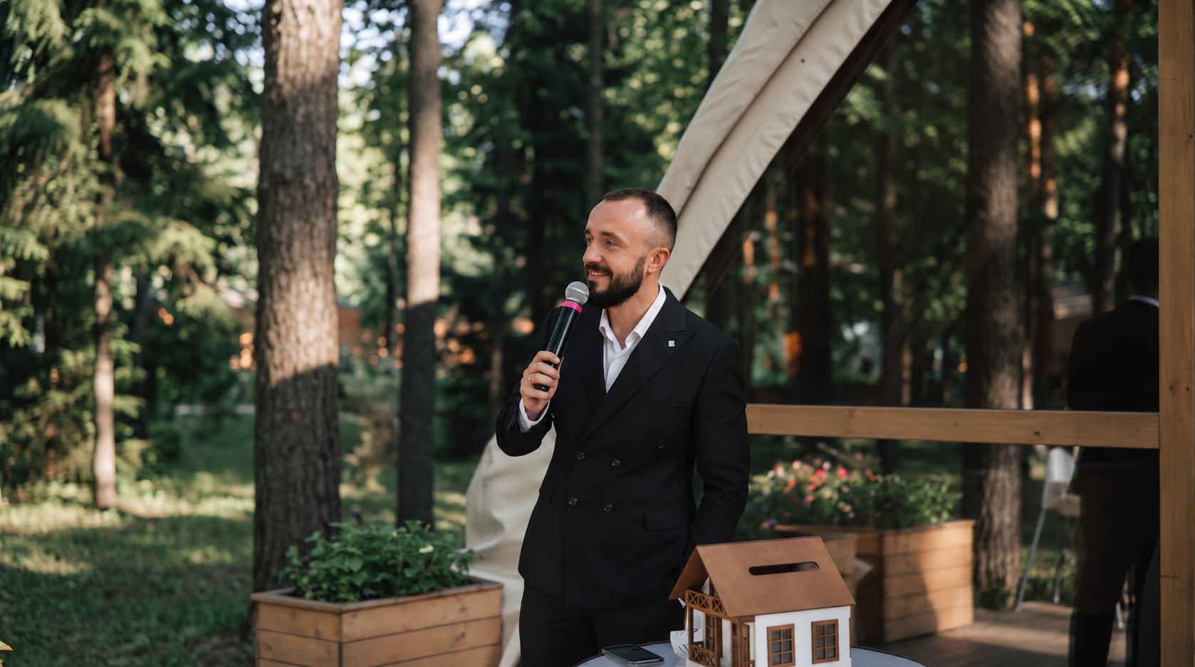 Jens lächelt mit Mikrofon im schwarzen Anzug vor Bäumen und einer verglasten Holzterrasse.