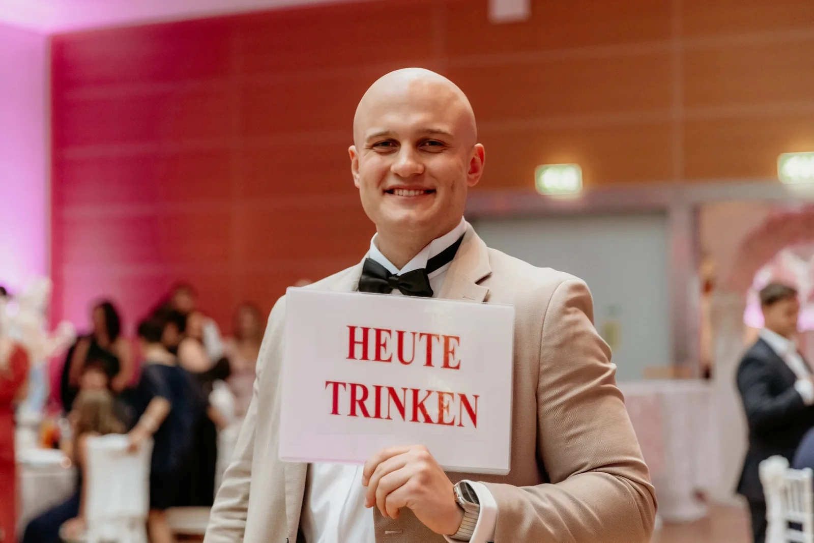 Tamada Eugen moderiert ein Hochzeitsspiel mit Schild „Heute trinken“ und sorgt für gute Stimmung.