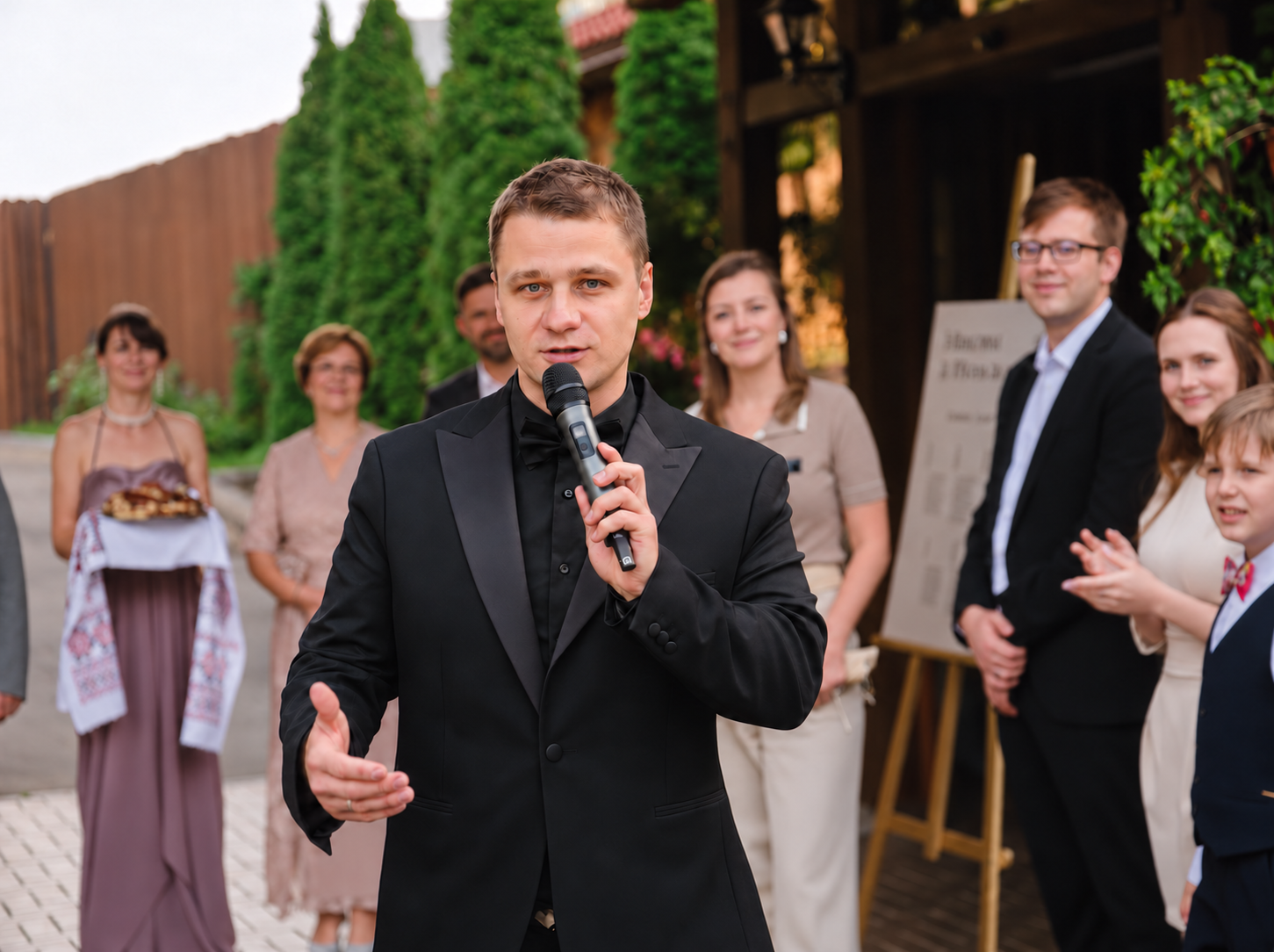 Dima Key im schwarzen Smoking spricht mit Mikrofon vor Hochzeitsgästen im Freien. Dima Key im schwarzen Smoking spricht mit Mikrofon vor Hochzeitsgästen im Freien.