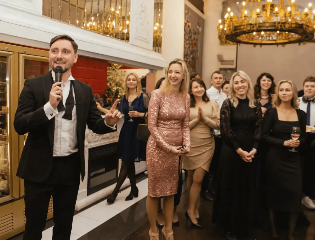 Sergey Solovyov moderiert mit Mikrofon inmitten festlich gekleideter Gäste.